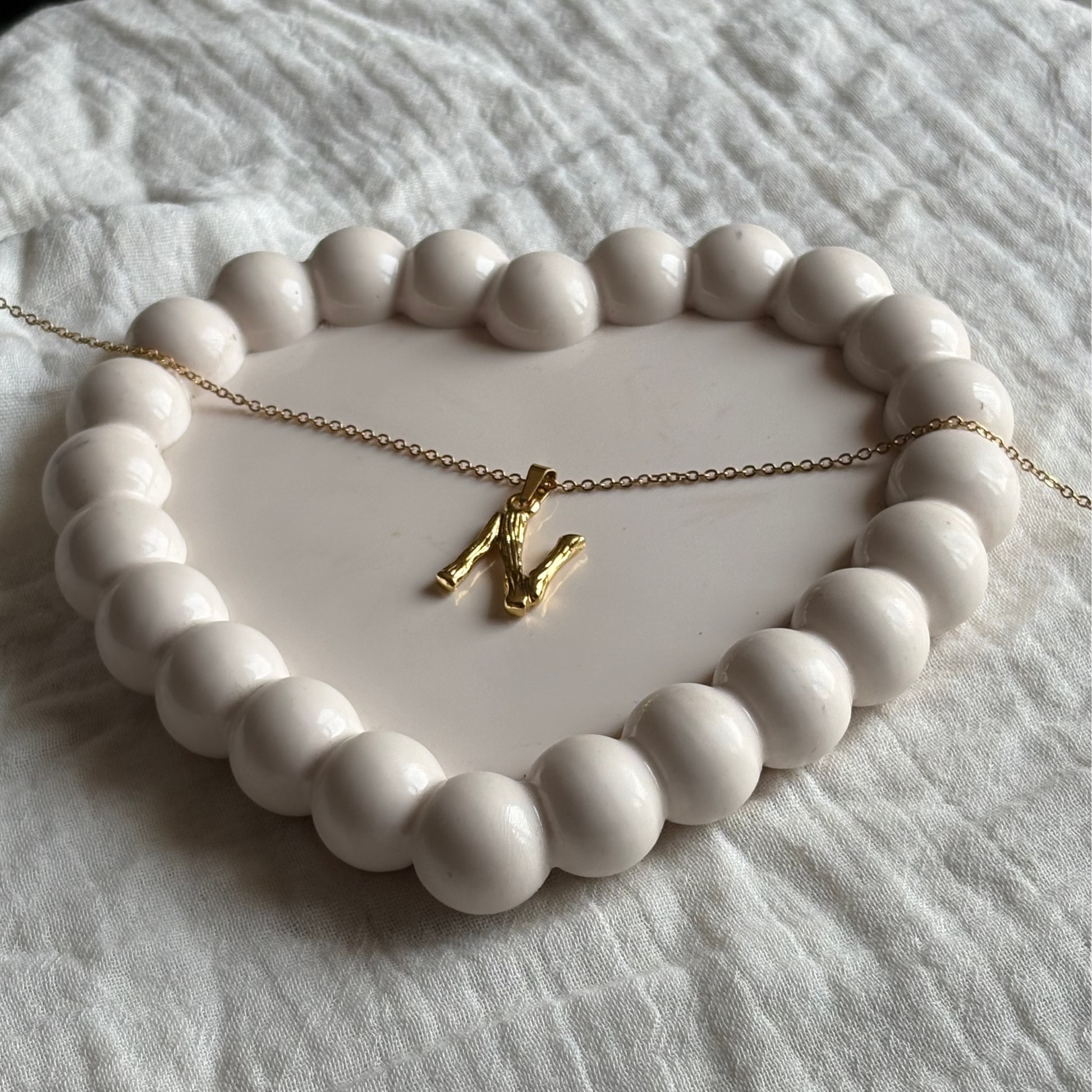 Gold necklace with a N shaped pendent on a white ceramic dish with a beaded edge.